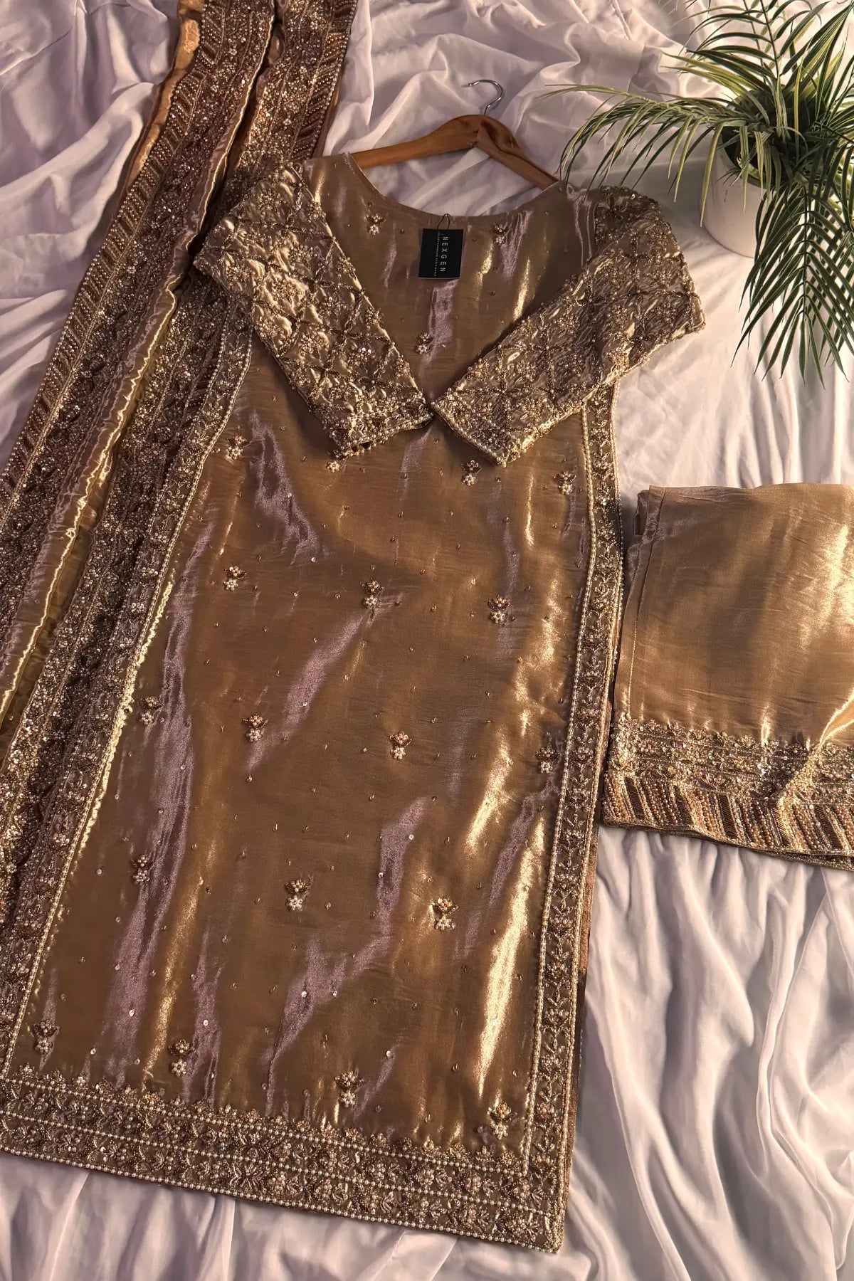 Gold embroidered traditional outfit on a white bed with a plant in the corner